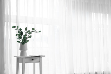 Vase with eucalyptus branches on table near light curtain
