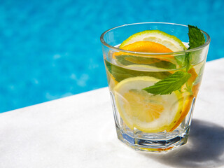 Citrus and mint leaves infused water, cocktail, lemonade in glass near pool. Summer iced cold drink with lemon, lime, orange, mint on blue water and white marble background, copy space, side view