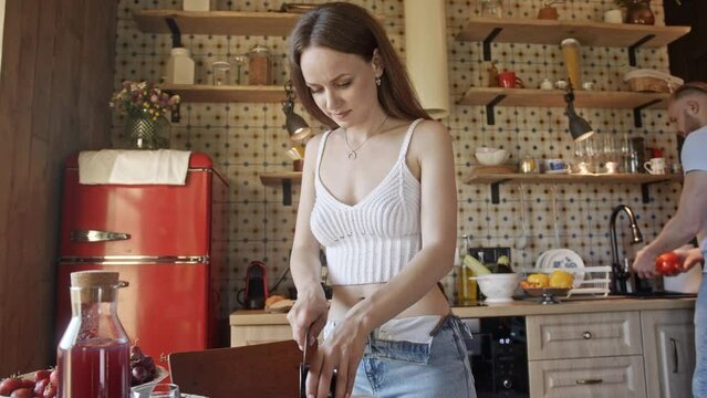 Lady cuts aubergine on wooden board putting pieces on plate. Woman cuts thin slices of aubergine and puts pieces on plate. Husband washes bell pepper and wife takes pepper from man in cozy stylish