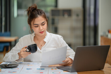 Businesswoman working with documents and drinking coffee in front of laptop computer at desk