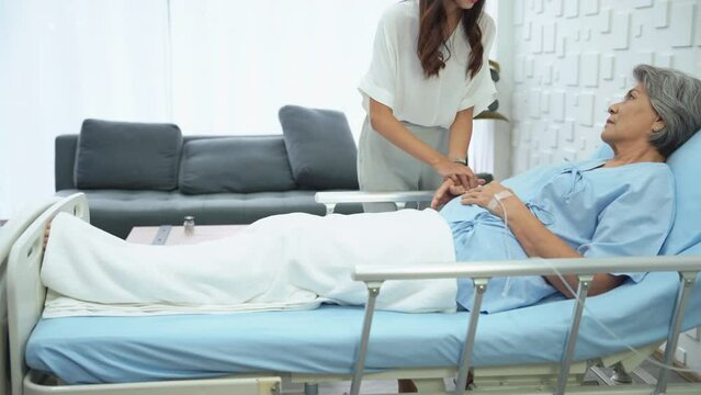 Young woman lifting a railing beside the bed and holding his sick mother's hand to get saline on the bed in the hospital room. Daughter encouraging mother lying sick in the patient room.