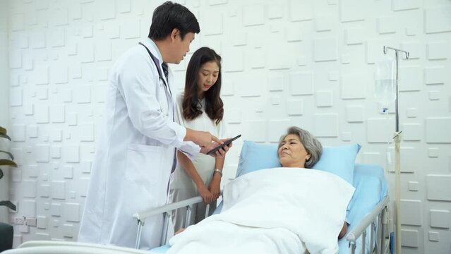 Specialist male doctor talking to the daughter of an elderly woman patient in the patient room about treatment programs and costs. Elderly female patient waiting in bed have a bright smile no worries.