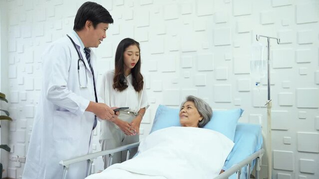 Specialist male doctor talking to the daughter of an elderly woman patient in the patient room about sickness and better treatment results. Elderly female patient in bed have a bright smile.