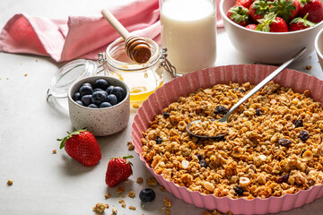 Homemade granola with raisin, seeds, hazelnut and peanut on baking sheet, milk or yogurt bottle, honey and blueberries, strawberries on grey background. Healthy breakfast ingredients. Selective focus.