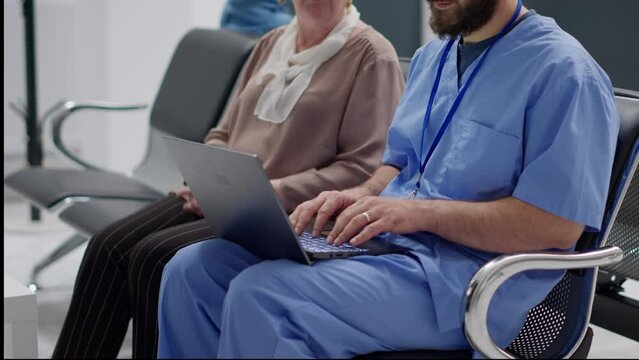 Medical assistant working on laptop to give diagnosis to old patient, consulting senior woman in waiting room. Male nurse and elderly person looking at computer to find medicine treatment.