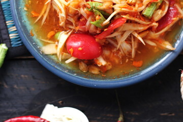 Spicy papaya salad served in a blue dish with sticky rice in a bucket and long beans, dried chilli and vegetables.