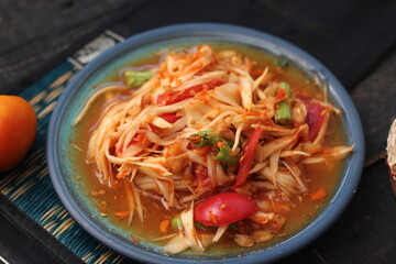 Spicy papaya salad served in a blue dish with sticky rice in a bucket and long beans, dried chilli and vegetables.