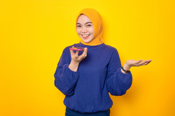 Cheerful young Asian Muslim woman dressed in casual sweater talking on smartphone using loudspeaker...