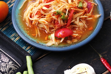 Spicy papaya salad served in a blue dish with sticky rice in a bucket and long beans, dried chilli and vegetables.