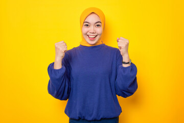 Excited young Asian Muslim woman dressed in casual sweater celebrating success with raised fist isolated over yellow background