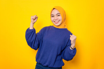 Excited young Asian Muslim woman dressed in casual sweater celebrating success with raised fist isolated over yellow background