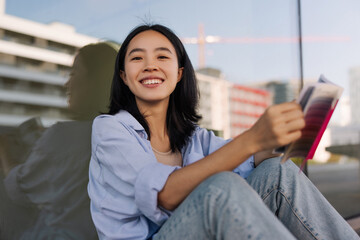 Brunette attractive chinese woman look with smile at camera sitting in the city with pink book. Wearing blue shirt, jeans. Concept of street style, student life