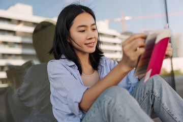 Charming young asian woman look and holding pink book on the street . Dark-haired pretty lady sitting on the floor ad reading. Relaxing, lifestyle 