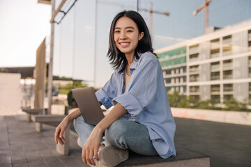 Side view of asian cheerful woman look with smile at camera in the city . Young student working outdoors sitting and using laptop. Technology, student lifestyle concept 