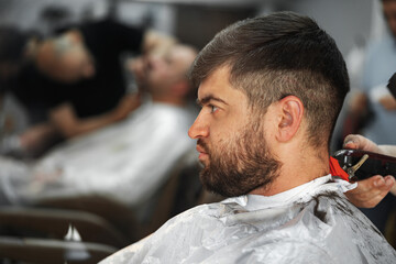 Barber guy gives a haircut to a bearded man sitting in a chair in a barbershop