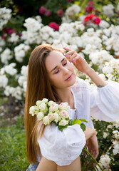 portrait of a beautiful brunette smiling woman in white embroidered shirt and jeans shorts with bouquet of white roses flowers  in summer park