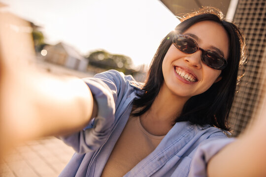 Happy Charming Brunette Woman Reach Hand To Camera And Smiling . Chinese Lady Wearing Blue Shirt In Sunglasses On The Street . Emotional, Technology Concept 