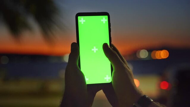 Close up smartphone with chroma key green screen at sunset dusk in male hands over tropical twilight, scrolling through social media or online shop - internet, communications concept 4k 