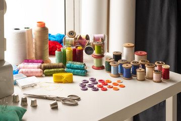 work table with spools of thread or bobbins. next to it a pair of scissors and a tape measure, in the background a sewing machine
