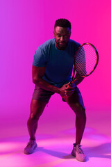 Vertical image of happy african american male tennis player in violet and pink neon lighting