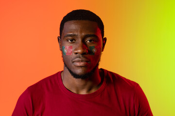 Portrait of african american male supporter with flag of portugal on cheeks over orange lighting