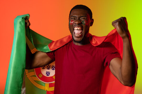 Portrait Of African American Male Football Supporter With Flag Of Portugal Over Orange Lighting