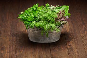 a bunch of green dill, parsley, salad and other greens in an iron bucket, dark wooden background, concept of fresh vegetables and healthy food