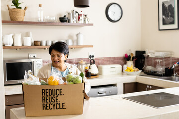 Biracial mature woman with short hair unpacking groceries from recycling box on kitchen island