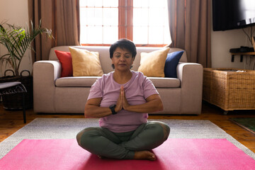 Full length of biracial mature woman meditating in prayer position on mat in living room at home