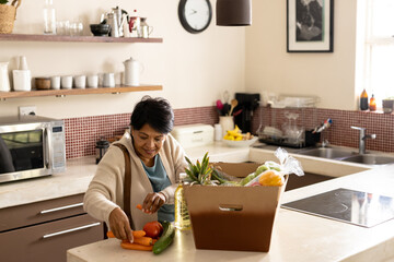 Biracial mature woman unpacking fresh vegetables and fruits from cardboard box on kitchen island