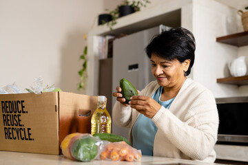 Biracial mature woman unpacking fresh vegetables and fruits from recycling box on kitchen island