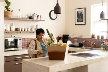 Biracial mature woman with short hair unpacking fresh groceries from cardboard box on kitchen island