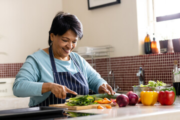 Smiling biracial mature woman with short hair wearing apron chopping vegetables on board in kitchen