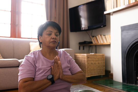 Biracial mature woman with short hair closing eyes and meditating in prayer position at living room
