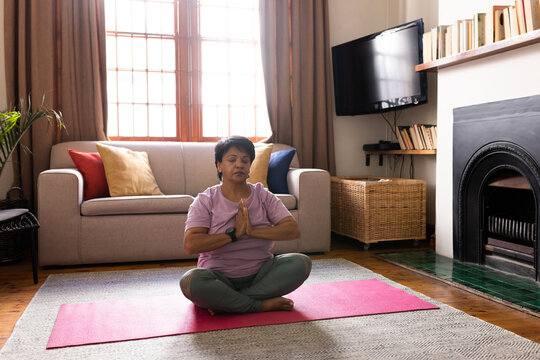 Biracial mature woman with short hair closing eyes and meditating in prayer position on mat at home