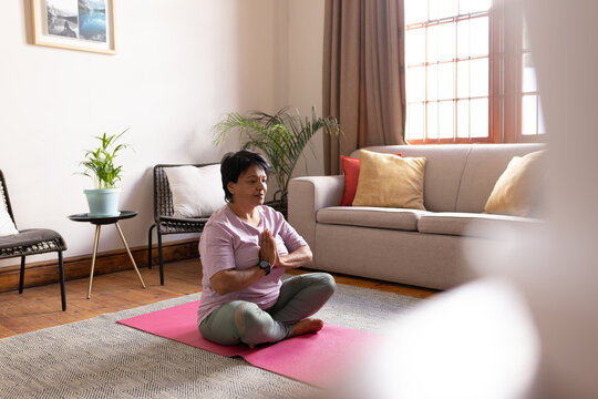 Biracial mature woman with short hair meditating in prayer pose while sitting on sofa in living room