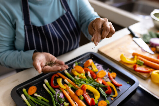 High angle midsection of biracial mature woman sprinkling spices on vegetables in tray in kitchen