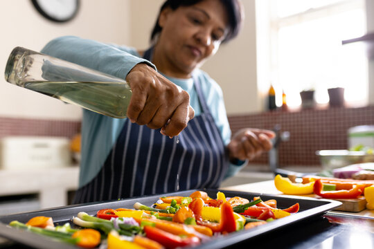 Biracial mature woman wearing apron spraying oil on fresh vegetables in tray in kitchen