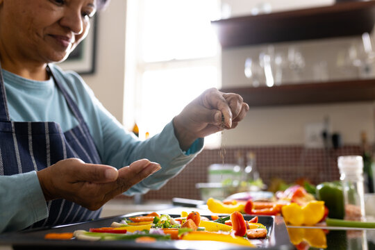 Midsection of biracial mature woman wearing apron sprinkling spices on vegetables in tray in kitchen