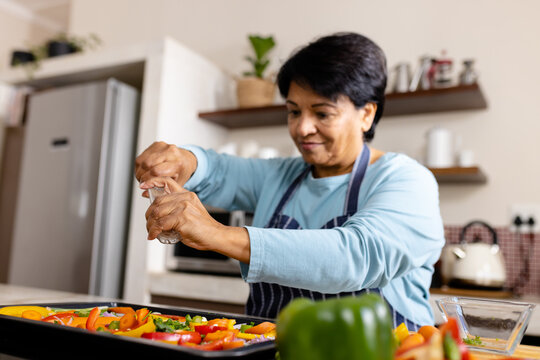 Biracial mature woman with short hair sprinkling pepper with shaker on vegetables in tray in kitchen