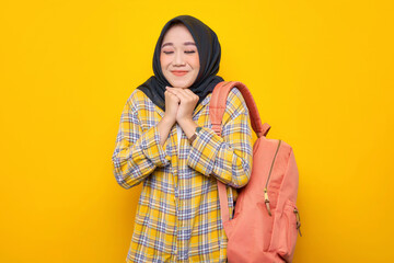 Fototapeta premium Smiling young Asian Muslim woman student in casual clothes and backpack, making a praying gesture with closed eyes isolated on yellow background. Education school university college concept