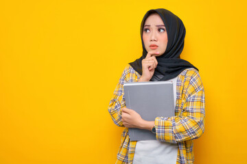 Fototapeta premium Pensive Young Asian Muslim student holding book, looks seriously thinking about a question isolated on yellow background. Back to school in high school college concept