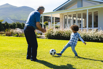 Caucasian grandfather playing soccer with biracial grandson on grassy land in yard on sunny day - Powered by Adobe