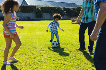 Multiracial multigeneration family playing soccer on grassy field in yard during sunny day