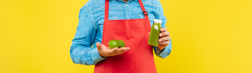 Man crop view in red apron holding limes and juice bottle yellow background, juicery