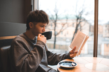 Young handsome teenage boy 18-19 year old drinking coffee sitting in cafe and reading paper book. Millennials lifestyle.