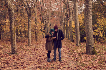 Real young white Caucasian boy and girl couple looking at smartphone in a park with brown leaves fallen on the ground in autumn