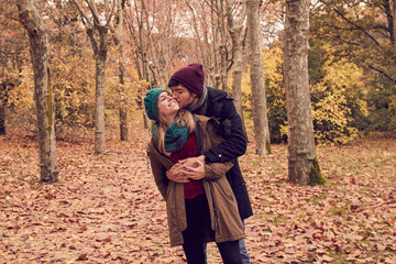 White Caucasian man hugging his girlfriend from behind giving her a kiss on the cheek while she is smiling in a park in autumn with the ground full of brown fallen leaves - obrazy, fototapety, plakaty