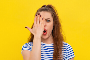 Close up portrait of shocked brunette girl, cover half of face and gasping startled, looking at camera with one eye, checking vision, standing in white-blue striped t shirt over yellow background