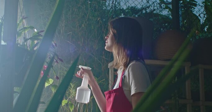 Young woman spraying water into plants inside greenhouse. Employee of small business caring for plants and flowers irrigating. Girl wearing apron watering plants